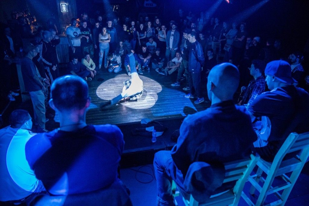 Breakdancer in a spotlight surrounded by a circle of spectators in blue-lit venue